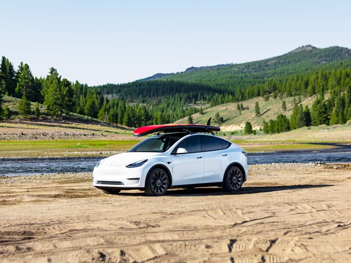 A white Tesla Model Y sedan carrying a red surfboard on its roof with mountainous terrain in background.