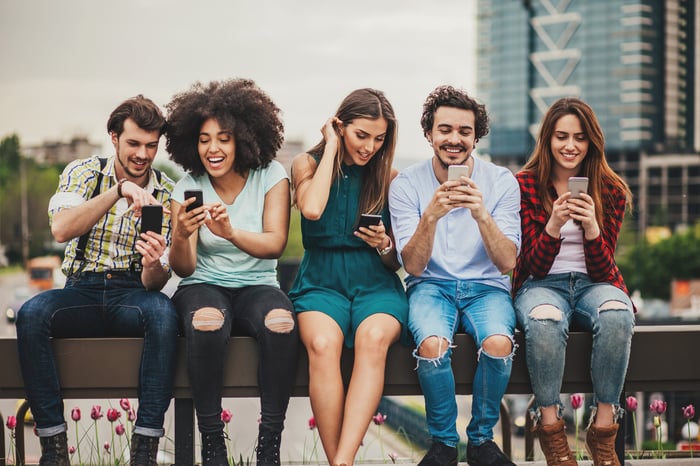 A number of young people sitting on a bench laughing and looking at their smartphones.